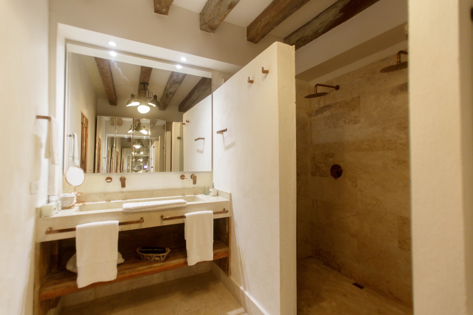 Casa Palmas bathroom with double travertine vanity, bronze fixtures, and open-plan rain shower alcove under colonial ceiling beams