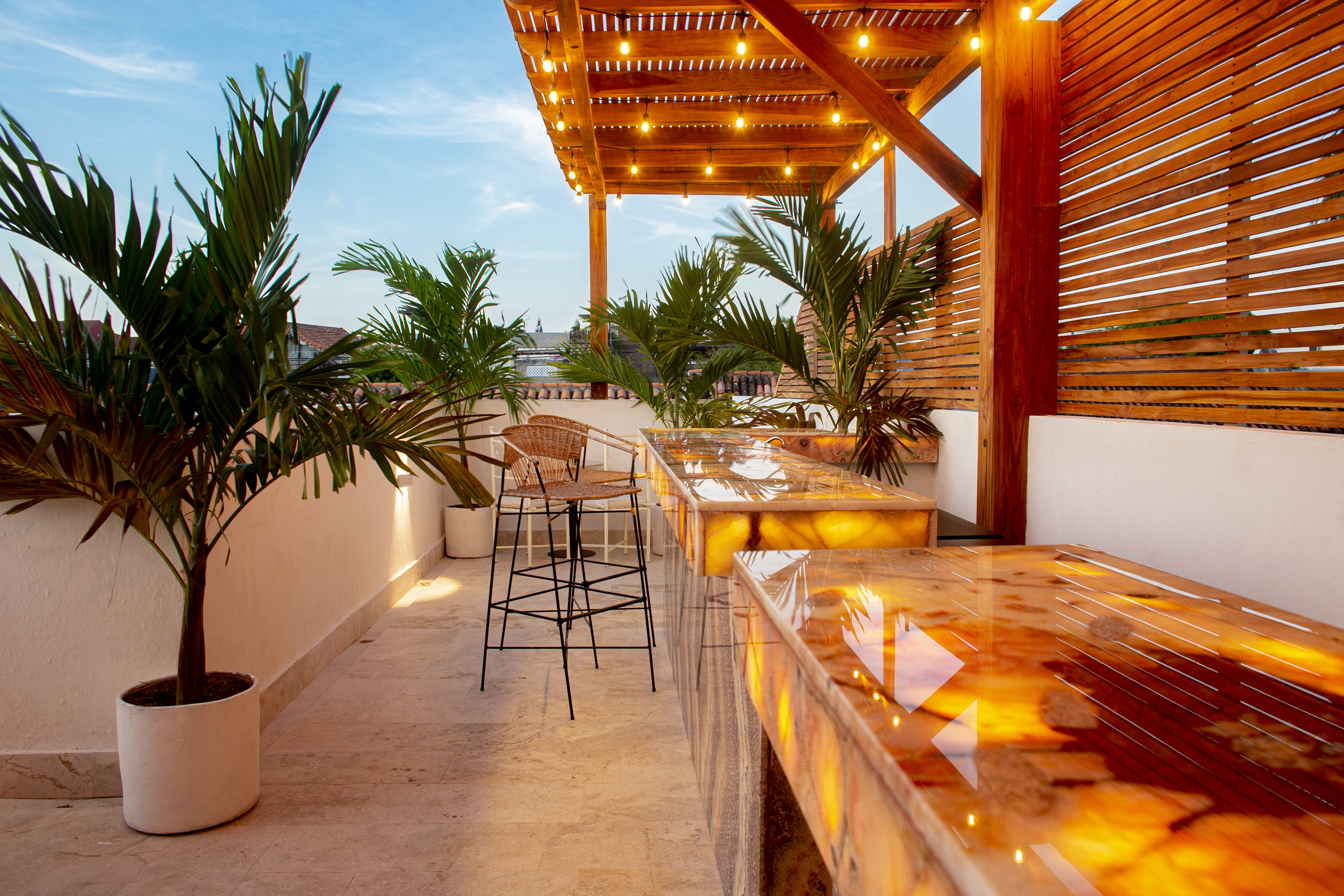 Casa Palmas rooftop terrace at sunset with two illuminated onyx bar counters, rattan stools, and string-lit pergola